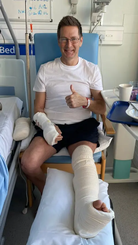 Submitted Jerry Innesbeer is sitting in a blue hospital chair next to a bed. He is wearing a white top and black shorts and his left leg is bandaged up. His right arm is also bandaged and his left has a bandage around his wrist. He is wearing rectangular glasses and has short brown hair which is slightly spiked up and he is pulling a thumbs up.