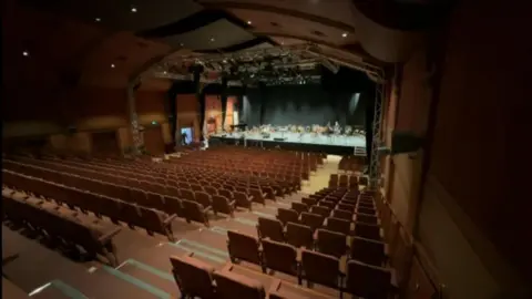 A view from the top of an auditorium. There are hundreds of seats in rows, separated by an aisle. At the bottom there is a stage, where there are seats set out for an orchestra or band.