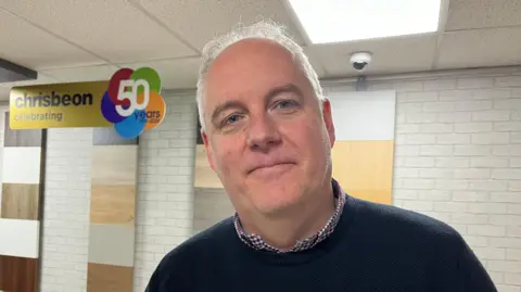 Chris is looking into the camera and smiling slightly; he's wearing a navy jumper with a red, blue, and white checked shirt underneath. He's stood in front of a whitewashed brick wall at his company that's got twelve different wood samples hung up. To his right there's a yellow sign with black writing that reads 'Chrisbeon - celebrating 50 years.'