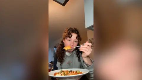 Rhi sits with a bowl of mango curry in front of her, she holds a spoonful up to the camera