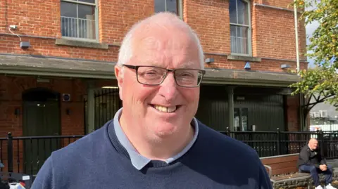 A man with white hair and dark rimmed glasses smiles into the camera. He is wearing navy jumper and light blue polo shirt. In the background a man in a black jacket is sitting on a bench. There is a red brick building in the background with black railings.  