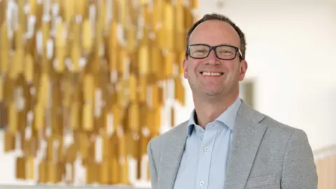 Gloucestershire Hospitals NHS Trust Richard is smiling to camera in a mid-shot. He is wearing black-rimmed glasses and a grey suit jacket with a light blue shirt. In the background is a blurred golden chandelier, which looks like it could be hanging in the high-ceilinged foyer of a hospital