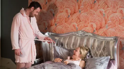 Danny Dyer as Mick in EastEnders, standing in a short pink dressing gown, extending the pink belt out as he smiles down at his EastEnders wife, Linda. Linda is under the duvet, laughing. There is a silver headboard and flamingo wallpaper behind them. 