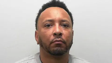 A custody photo of a man wearing a light grey sweatshirt who sits against a plain, pale background, facing forward.