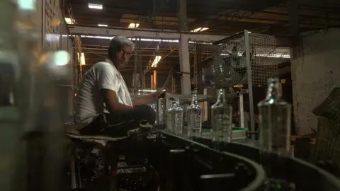 A worker - wearing a white undervest and a cotton scarf around his neck - sits next to a conveyer belt carrying glass bottles in a factory