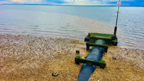 STEPHEN HUNTLEY/BBC A large pipe extends across a pebbled beach into the sea, with calm water and a distant shoreline visible under a cloudy sky. A stream of brown water is emanating from the pipe into the blue sea.