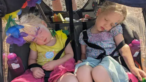 Family handout Vicky's two daughters fast asleep in a big buggy. They both have festival face paints, with floral and bright coloured dresses. There are multi-coloured flowers adorning the buggy.