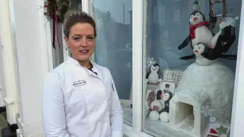 A woman with dark hair pulled back from her face wears a chef's whites. She is standing next to her cake shop window display, which features a large, iced igloo and cake penguins.