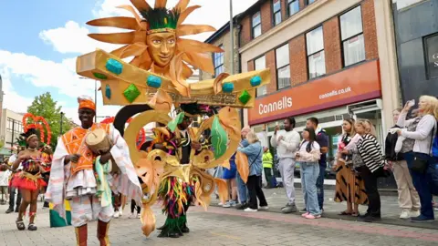 Stephen Huntley/BBC A person wearing a giant elaborate gold costume, with a large face on top, is part of the parade at Luton Carnival. Onlookers are watching for the side. A man with a drum is walking alongside it.