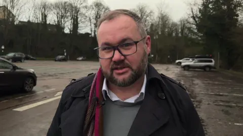Nicola Rees/BBC A picture of Councillor Alan Lamb with glasses and a beard stand in Wilderness Car Park in Wetherby with some cars and trees in the background but lots of empty parking spaces.