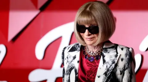 Anna Wintour, in her signature black sunglasses, poses on a red carpet in a red top and black-and-white patterned jacket