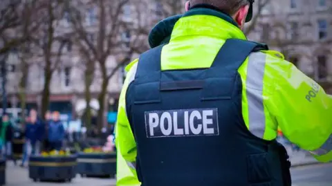 A PSNI officer is standing with his back to the camera wearing a high visibility coat and bulletproof vest. He is on patrol in Belfast city centre as pedestrians walk by.