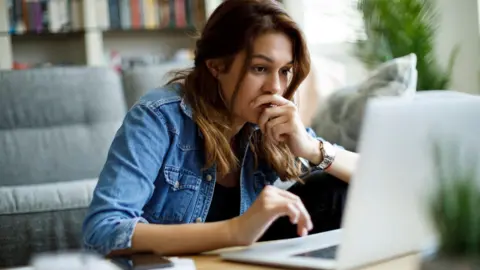 Getty Images Wanita muda dengan rambut coklat, mengenakan jaket denim, melihat layar laptop