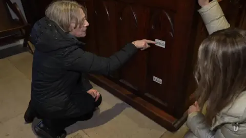 Sarah Lunn, a vicar dressed all in black with shoulder length brown hair and glasses points to a sticker on the end of pew which shows where flood water got to in 2015. About a foot below it is another sticker from 2009, which is also about a foot off the grouind