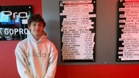 Daytona Milton Keynes Kimi Antonelli smiling, standing by a celebrity leader board, with lots of names on it. He is wearing a white top, with a hood, smiling at the camera and to the left of the image. 