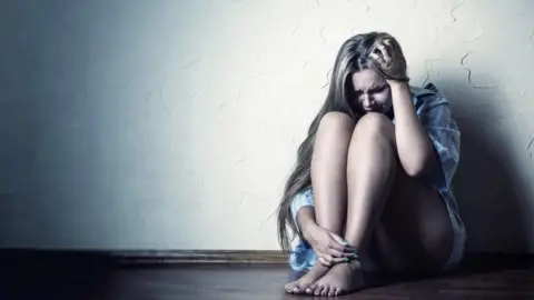 Getty Images A woman sits in the corner of a room on the floor with her head in her hand. She looks distressed.