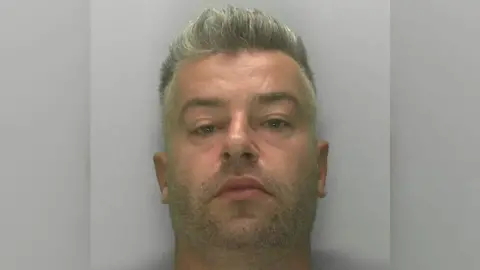 Gloucestershire Police Artur Mieszczak pictured in a custody photo, against a grey background. He has grey hair gelled upwards and is looking blankly at the camera. 