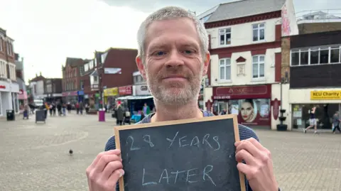 Kate Bradbrook/BBC A man holding up a board which reads "28 Years Later". He is standing in Kettering town centre