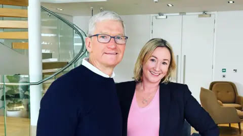 Tim Cook wearing a dark blue sweater, posing for a photo with Zoe Kleinman, wearing a pink top and dark jacket