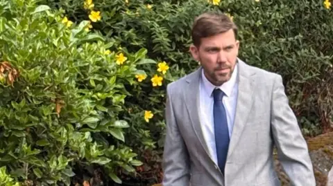 Lewis Baker has dark hair and a dark beard and is walking in front of green bushes. He is wearing a grey jacket, white shirt and blue tie.