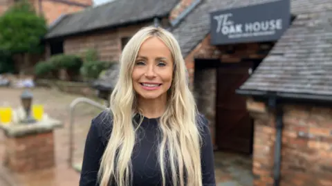 A smiling woman with long blonde hair and wearing a black top stands in front of a building with a sign over the entrance that says "The Oak House". 