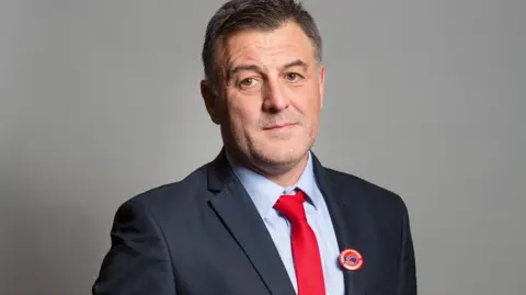 Ian Byrne, who has dark hair greying at the sides, poses for a picture wearing a navy blue suit with a red tie. 