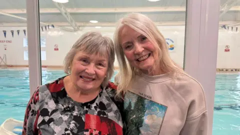 BBC The photo shows two women smiling at the camera. The lady on the left has short hair, and is wearing a floral top. The lady on the right also has short hair, and is wearing a pink jumper. They're standing in front of a window. There is a swimming pool behind them. 