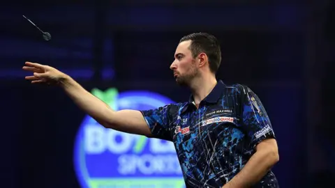Getty Images Darts player Luke Humphries on stage wearing a blue and black collared t-shirt and throwing a dart.