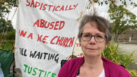 BBC Jenny Read is sitting on a camping chair protesting outside the Church of England's parliament, the General Synod. Behind her is a banner which reads 'sadistically abused in a NE church, still waiting for justice' - although the word "for" is hidden in the fold of the banner. It's written in red, green and black on what looks like embossed, cream furnishing or curtain fabric.