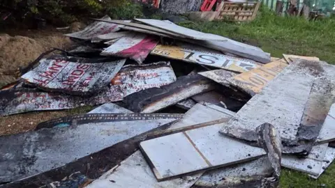 Close up photos of burned out advertising hoardings in a pile. The boards are all blacked from burning. The hoardings are on grass. 