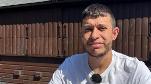 Arwel faces the camera, wearing a white t-shirt. He has short dark hair and stubble and is stood in front of a large wooden shed-type structure.