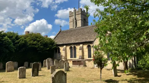 Shariqua Ahmed/BBC The churchyard with a cemetery in front - with tombstones and the historical church building in the background with a tower - surrounded by trees on either side.