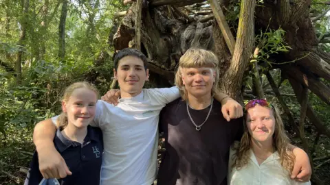 Four young people stand together in a woodland setting with their arms around each other. Behind them is a large treehouse-like structure built from branches and timber, surrounded by trees and dense greenery.