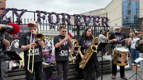 A brass band wearing black Ozzy Osbourne t-shirts stand in front of the Black Sabbath bench in Birmingham. A purple floral arrangement reading 'Ozzy Forever' is visible behind them.