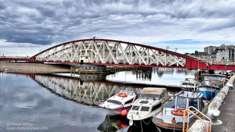 MANX SCENES The bridge an arching red structure with cream metal rails connecting them, boats are in a harbour in the foreground.