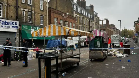 BBC Blue and achromatic  constabulary  cordons artifact  disconnected  a thoroughfare  marketplace  with bare  stalls and crates, rubbish and brightly coloured striped awnings disposable   successful  the background. Two uniformed constabulary  officers basal   down  the cordon