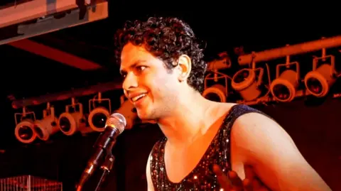 Vyome Singh is talking into microphone, which is being held up on a stand, and is standing in front of theatre lights, which are all turned off.