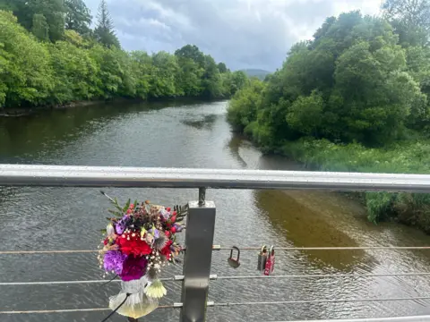 A bunch of flowers attached to a bridge over a river. There are full, green trees growing on the banks of the river. 