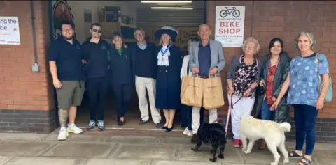 Lucia Bridgeman  There are 10 people pictured in front of the shop including the High Sheriff of Northumberland, Lucia Bridgeman, who is wearing a traditional hat with a feather. There are also two dogs. On the wall are signs saying Bike shop and Changing the Cycle.