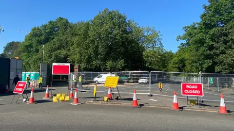 A road is fenced off at a junction with metal fencing and red-and-white cones. Two red signs with white writing say "Road Closed". Four security guards in high visibility yellow bibs are at the entrance to the festival site.