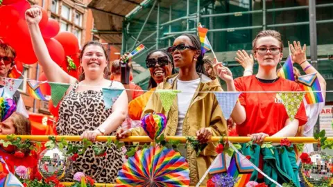 Getty Images A close-up of a group of people stand on a pride parade float, decorated with balloons, bunting, disco balls and rainbow garlands. Four people are prominent in the photo enjoying the festivities, raising their hands, waving flags and smiling.