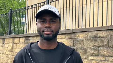 BBC/Grace Wood Daniel Obiku, wearing a black T-shirt and hoodie with a white baseball cap.