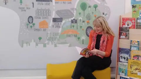 Artist Dora Colquhoun with long blond hair wearing an orange cardigan and black trousers and top sits on a yellow seat reading a book with a book case filled with books to the left. She sits in front of a wall with a map of Liverpool painted by an artist on the grey wall.  