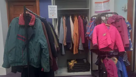 Several coats on a rack. It's on the side of the room in a library. There's a bright pink small coat on one side and a big green coat on the other side.