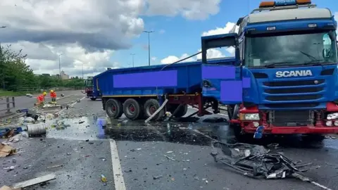 National Highways A blue-and-red Scania lorry with damage to its front has come to rest lying across all three lanes of a motorway. The road is covered in liquid and debris.