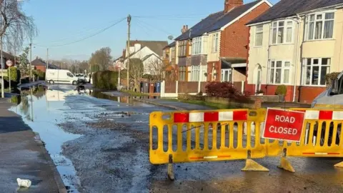 Image of water flooding sections of the road next to houses. Yellow barriers are placed in front with a red sign saying 'road closed'.