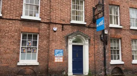 The outside of a brown brick building with a blue door.  It has a blue sign hanging above the door that says "The Hive", there is a similar sign on the left side of the door