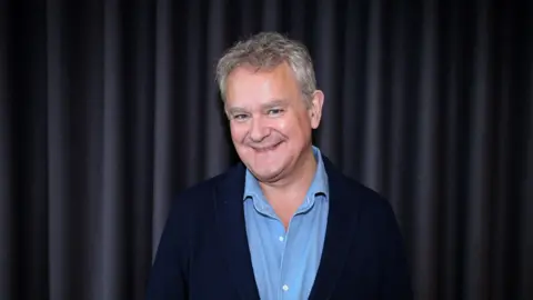 BBC The image shows a man smiling at the camera; he is wearing a light blue buttoned top and a navy blue blazer.
