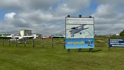 Dunkeswell Aerodrome. A sign for the airfield with a white aircraft on it next to a wooden perimeter fence. Several aircraft are on the other side of the fence.