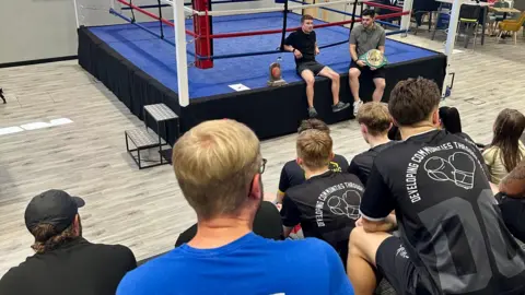 EMPIRE FIGHTING CHANCE A group of people on tiered seating with their backs to the camera look down at a boxing ring where two men sit in shorts and T-shirts speak to them.